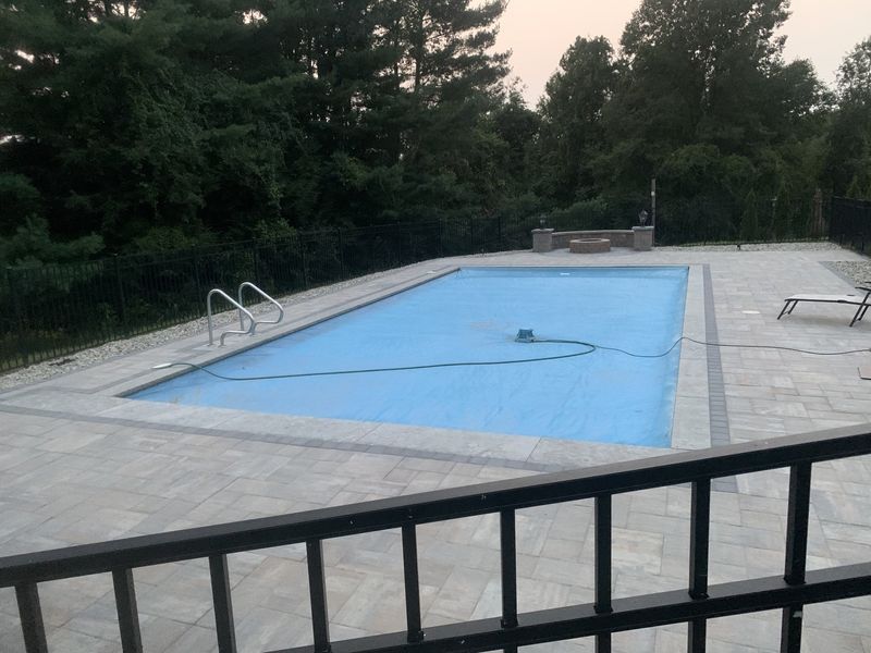 A rectangular swimming pool covered with a blue tarp, surrounded by a stone patio, black metal fencing, and trees.