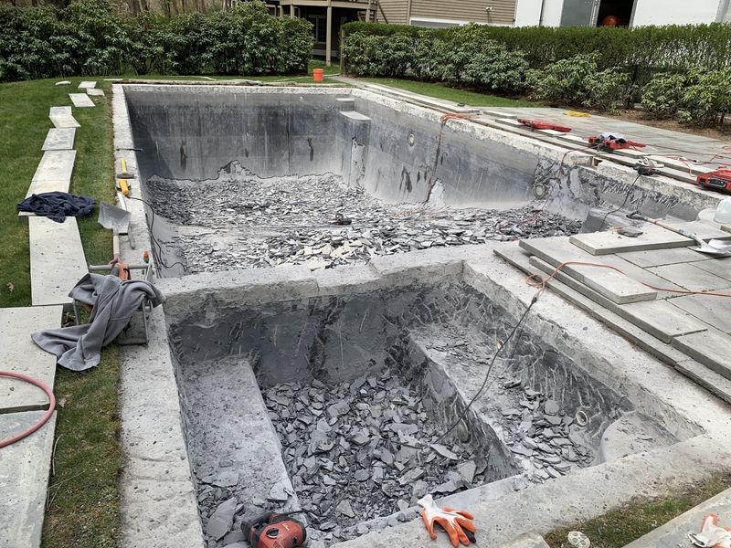 A swimming pool and attached hot tub being renovated with debris covering the bottom and surrounding stone coping.