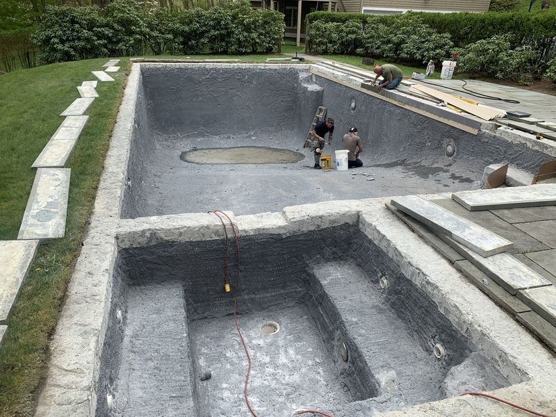 Workers apply a dark gray finish to the interior of a large empty swimming pool and attached hot tub in a backyard.