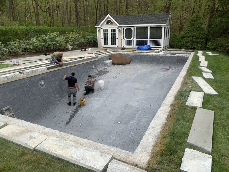 Workers install stonework around an empty, grey-lined rectangular swimming pool in a backyard with a small shed.