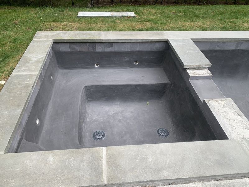 A dark grey concrete hot tub with built-in seating, set into a stone patio adjacent to an unfinished swimming pool.