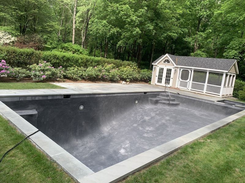 An empty, dark-bottomed swimming pool with stone coping next to a small screened-in pool house, surrounded by trees.