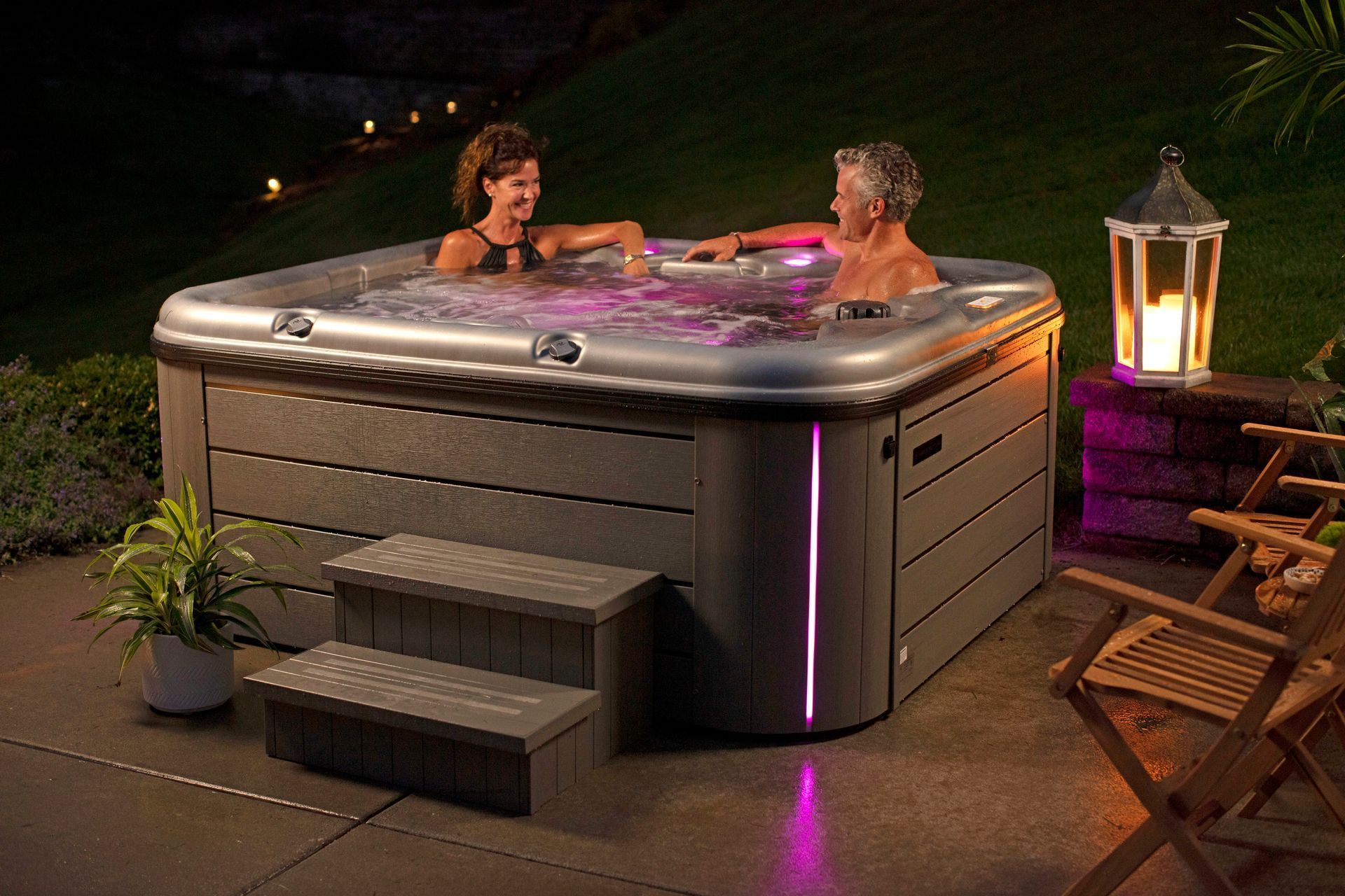 Two people relaxing in a glowing hot tub on a patio at night with a decorative lantern nearby.
