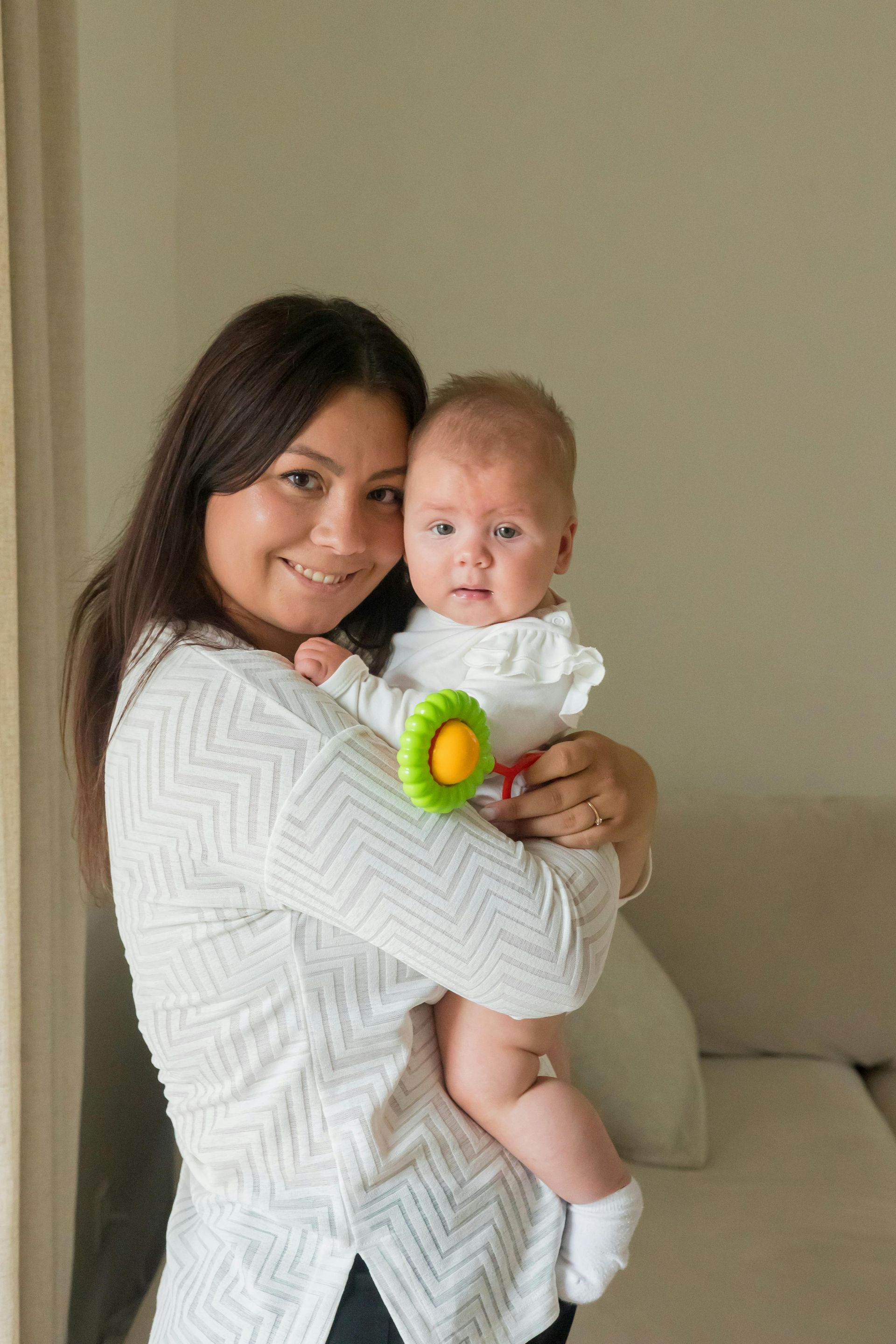 Woman holding a baby with a green toy in a beige living room