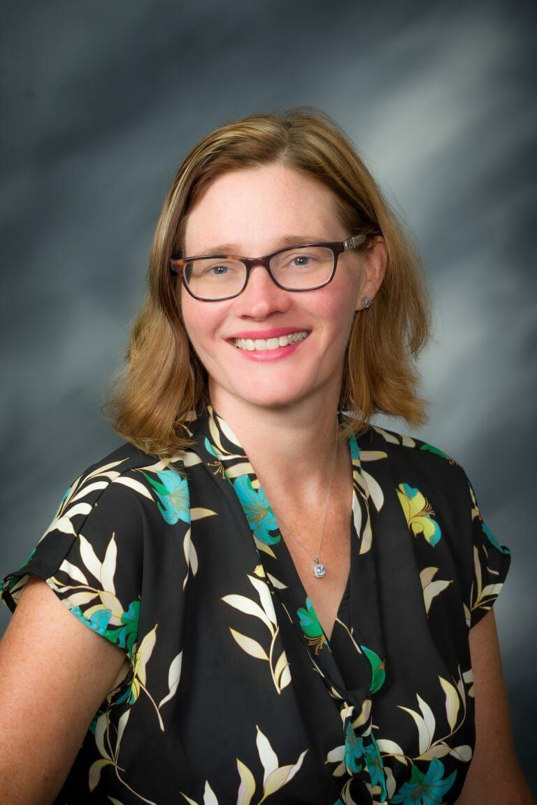 A woman wearing glasses and a purple shirt is smiling for the camera.