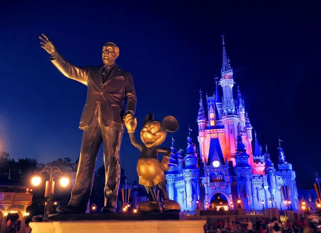 Statue of Walt Disney and Mickey Mouse in front of Cinderella's Castle at night, lit blue and pink.