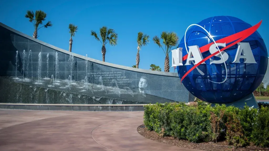 NASA logo with red swoosh, in front of a fountain and palm trees at the Kennedy Space Center.