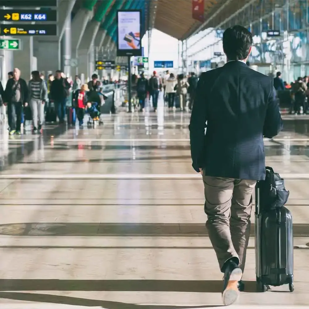 Man with rolling suitcase walking through a crowded airport terminal.