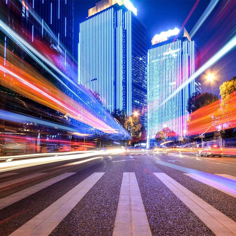 Nighttime cityscape with blurred streaks of light across a crosswalk and brightly lit skyscrapers.