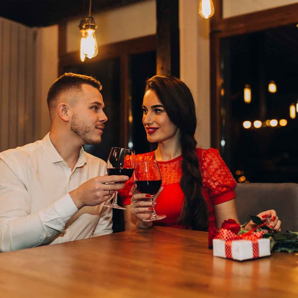 Couple toasting wine glasses at a restaurant table, he smiles at her, she smiles back. Gift box and roses present.
