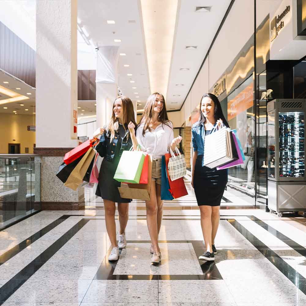 Three smiling women walking in a mall, holding shopping bags. Brightly lit interior with stores in the background.