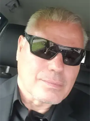 Man with fair skin wearing black sunglasses, suit, and sitting in a car.
