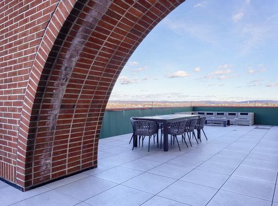 Brick archway frames a rooftop terrace with a table, chairs, and distant cityscape under a blue sky.