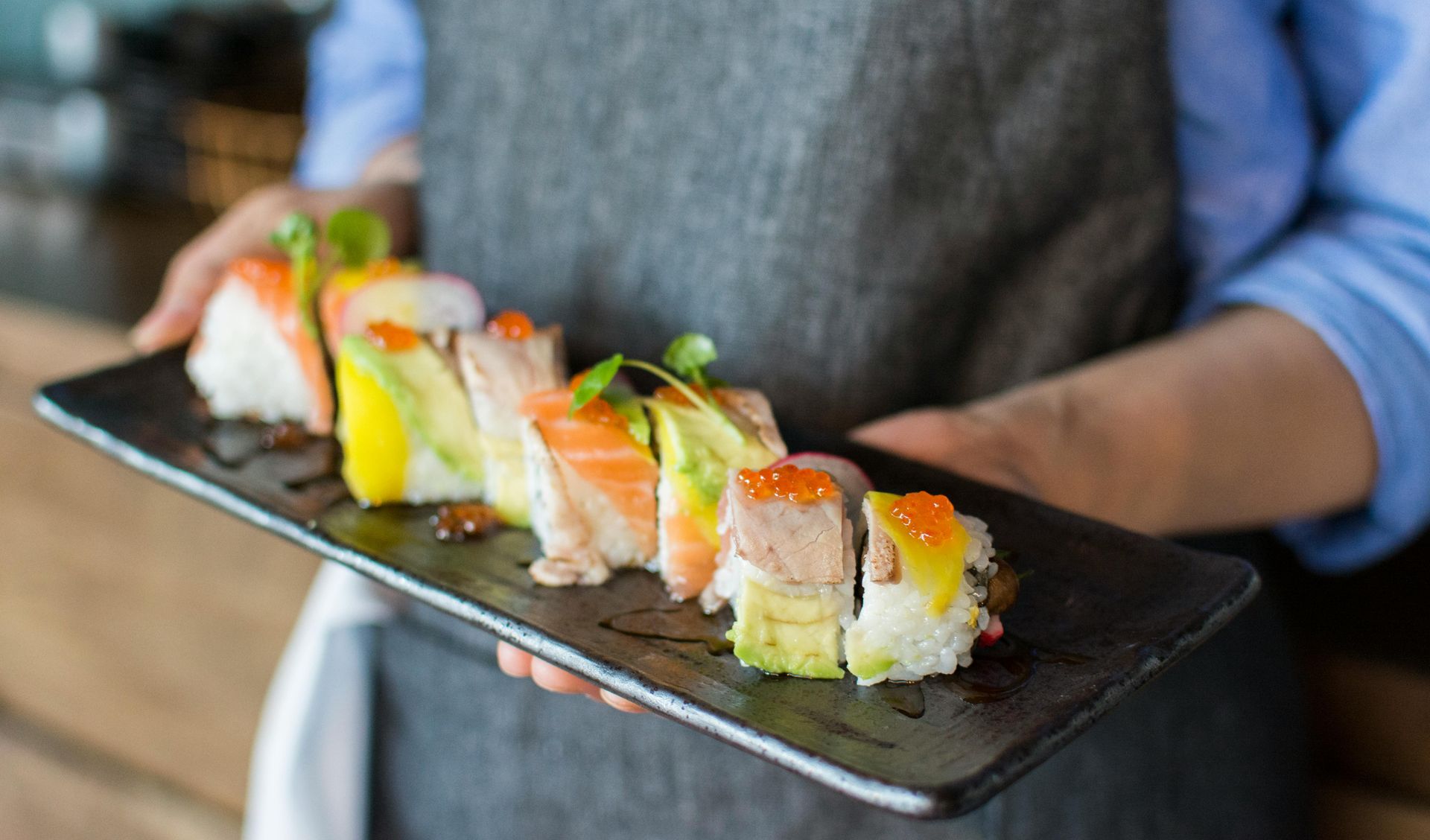 Sushi on a black plate held by a person wearing a gray apron and blue shirt.