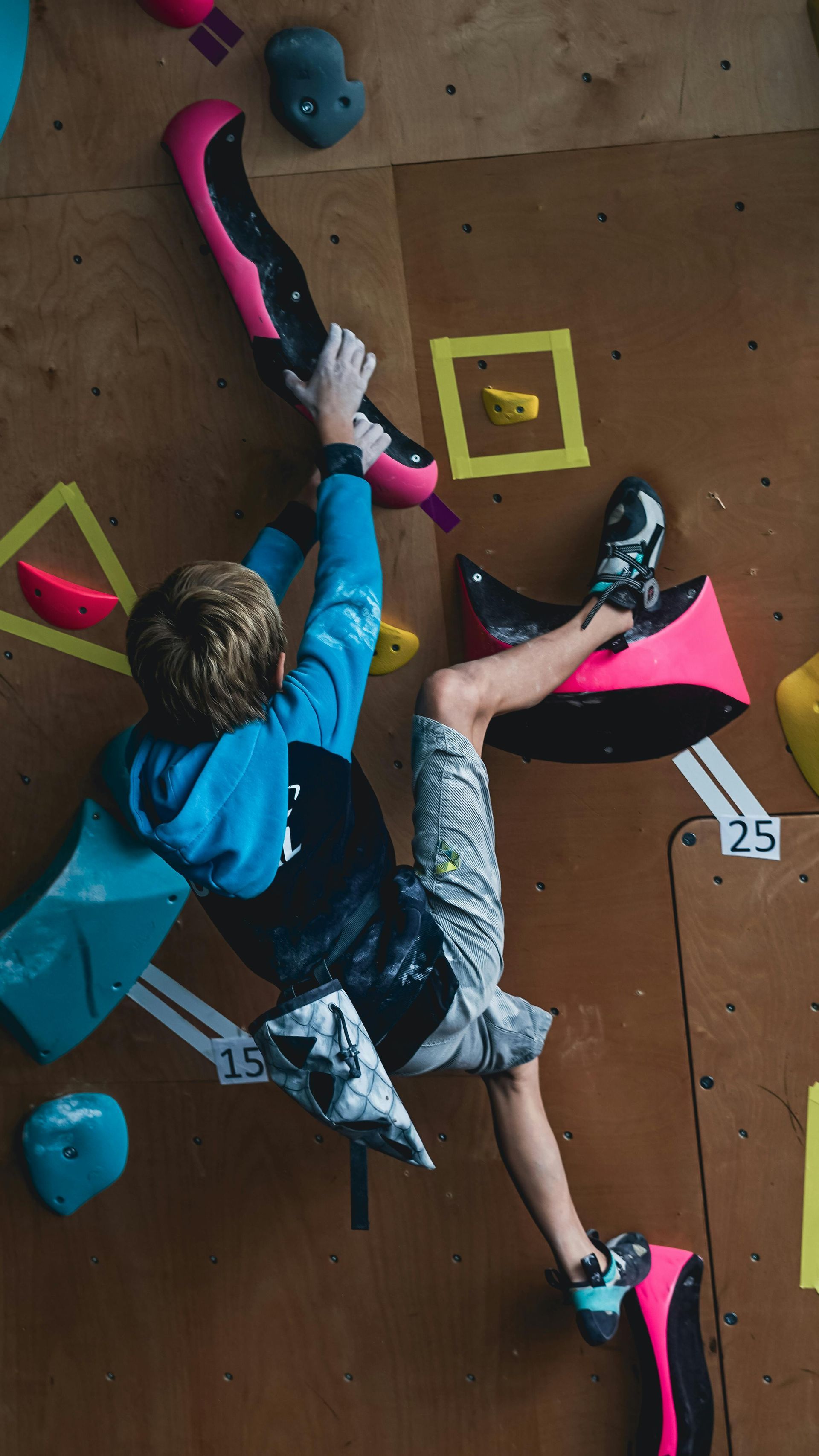 Boy climbing indoor rock wall, reaching for pink hold, wearing blue, black, and gray clothing.