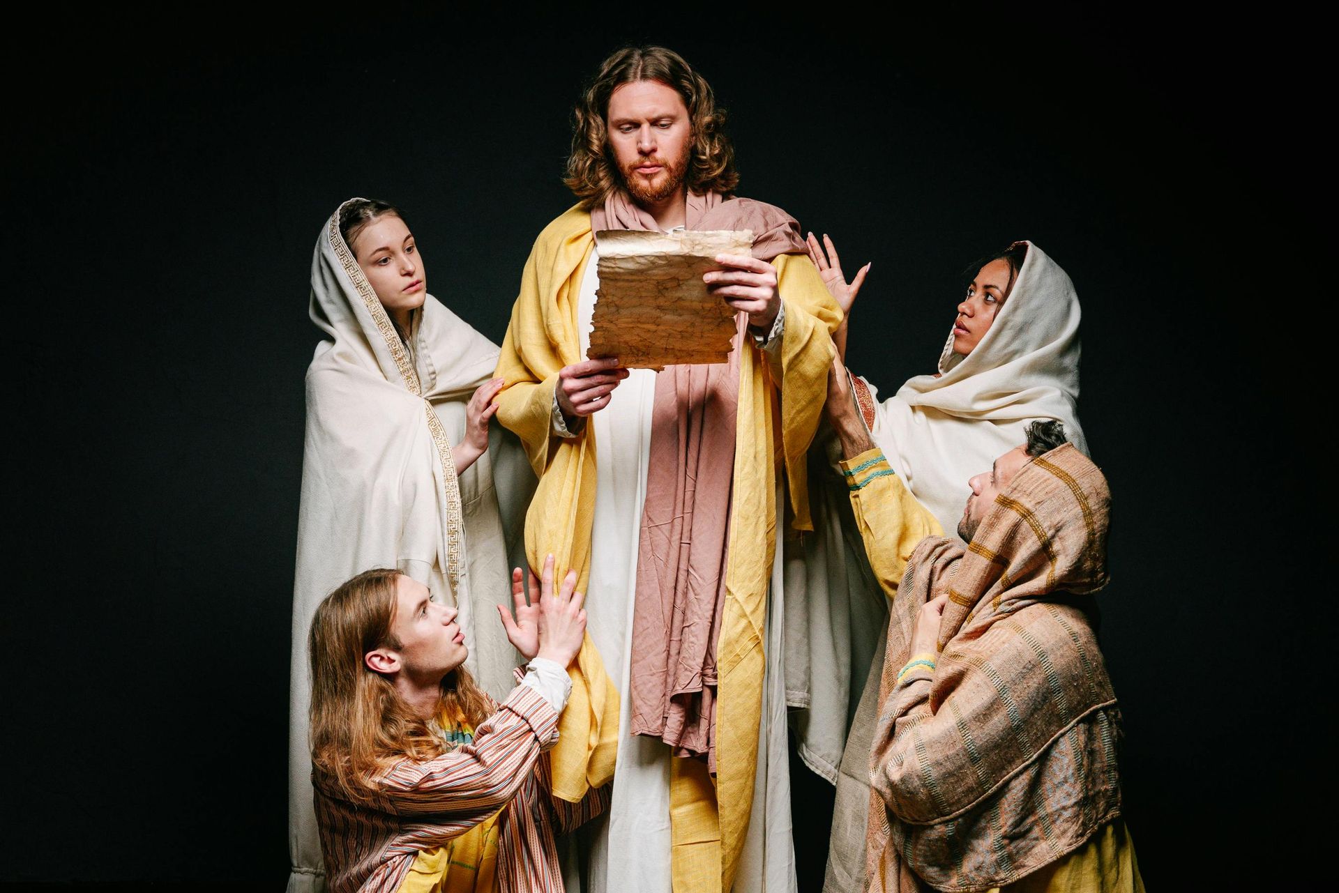 Jesus, in yellow robe, holds scroll as women in white and brown robes reach towards him. Dark background.