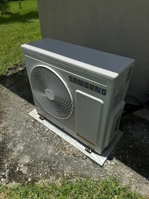 A Samsung air conditioning unit, white, is outdoors on a concrete pad, next to a white wall, with grass nearby.