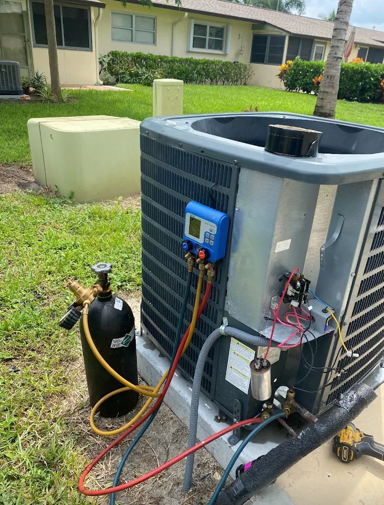 HVAC unit being serviced outdoors with gauges, hoses, and a refrigerant tank; residential setting.