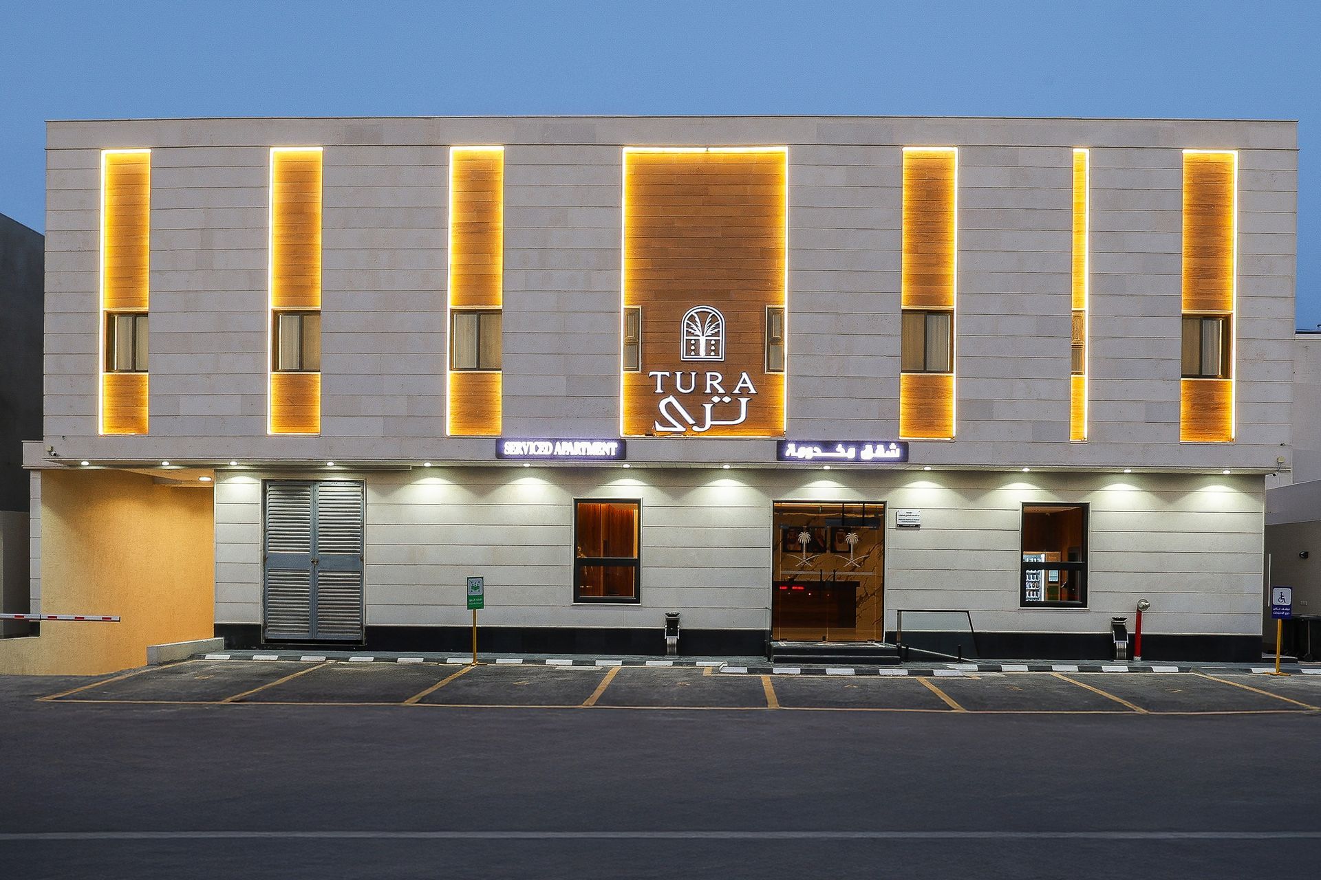 A modern, two-story stone building at dusk, featuring vertical golden accent lighting and the TURA logo centered.