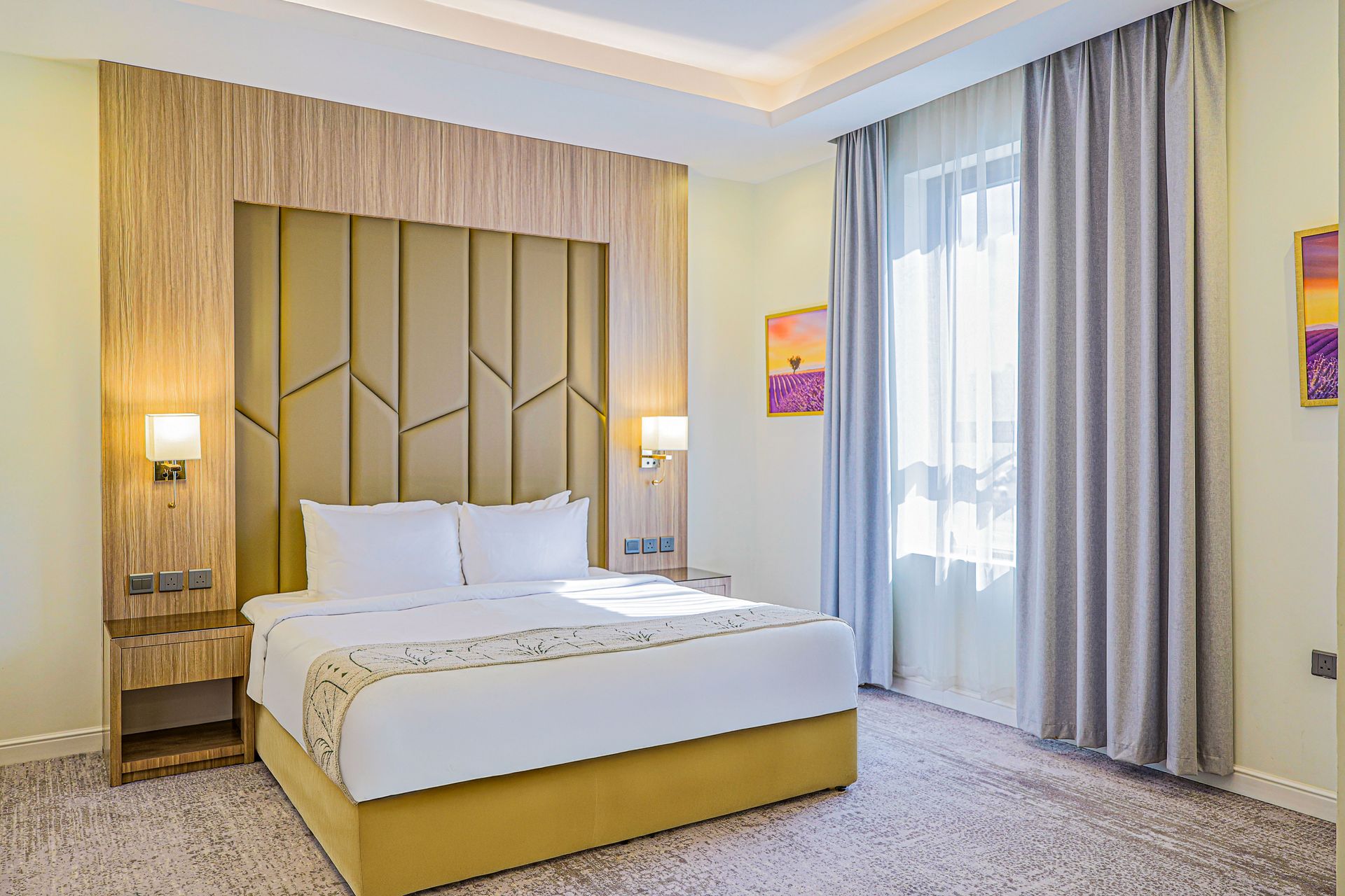 A modern hotel bedroom with a queen bed, a yellow tufted headboard, wood paneled accents, and gray floor-length curtains.