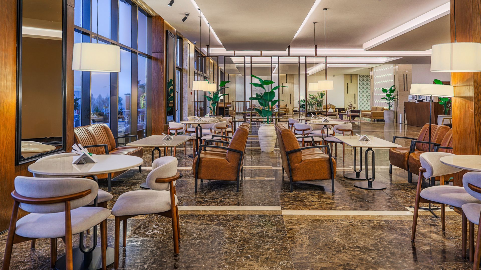 A modern, upscale hotel lounge with marble floors, leather armchairs, dining tables, and large windows with city views.