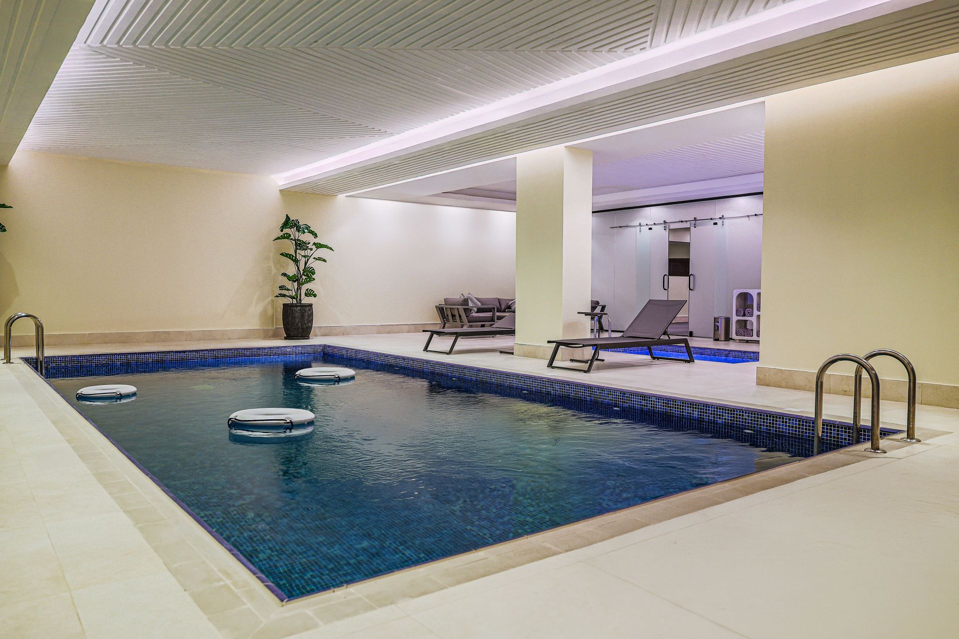 Indoor swimming pool with blue tiles, lounge chairs on the deck, and a modern ceiling with ambient lighting.