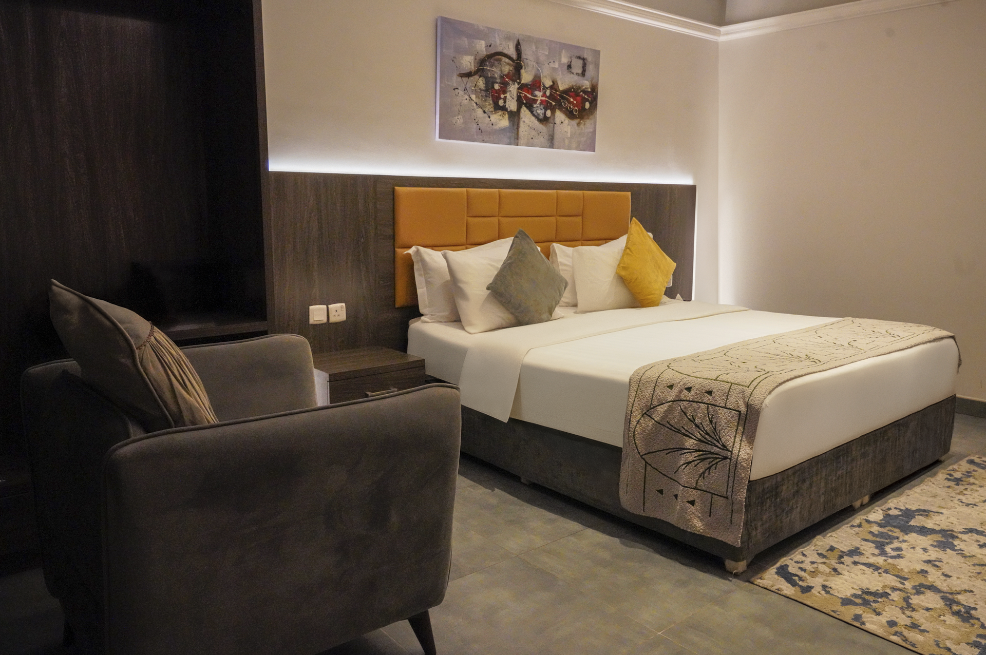 Modern hotel bedroom with a bed, armchair, dark curtains, and a patterned rug.