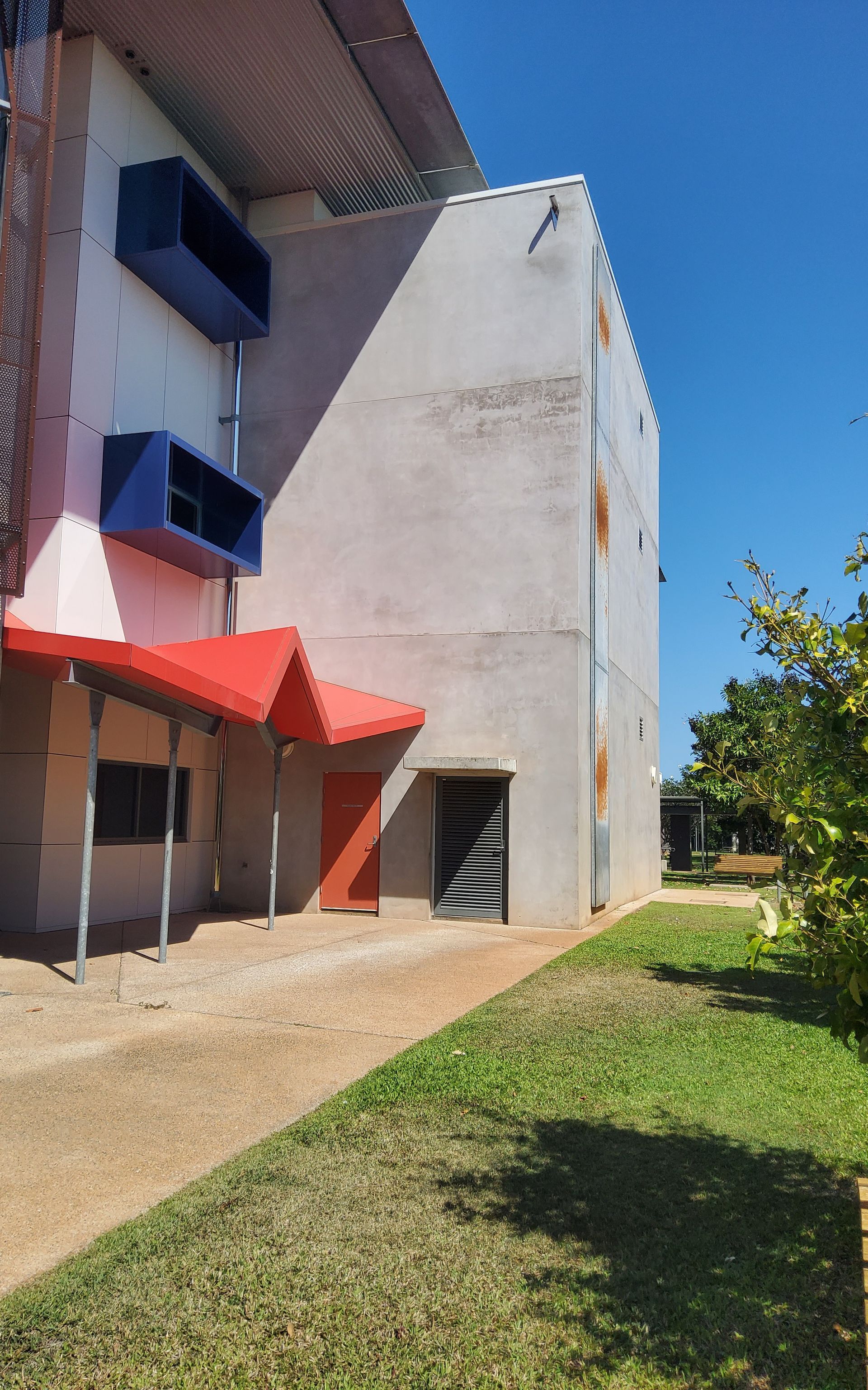 Side view of a building with blue air vents, red awning, and a path with grass on a sunny day   — Neil Griffiths Rendering in Berry Springs, NT