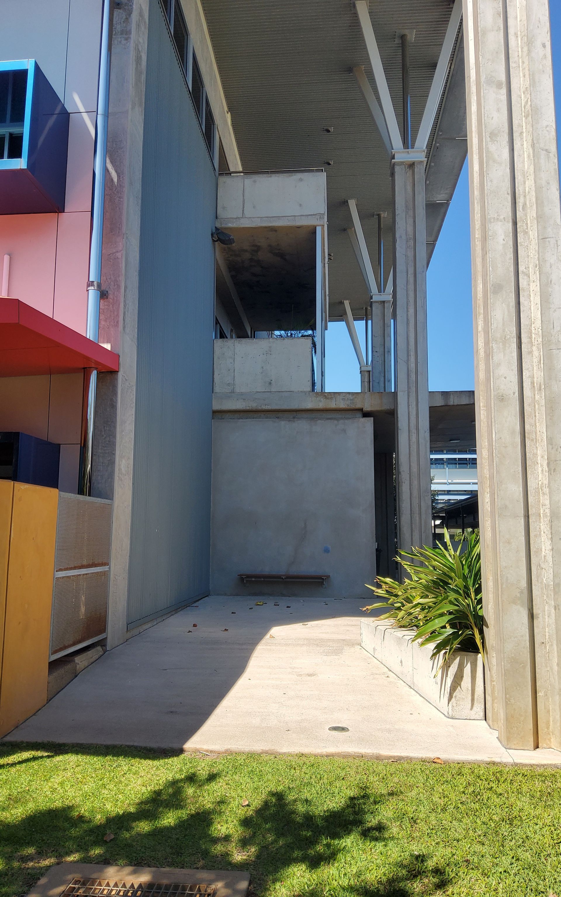 Ramp to gray wall with a bench, plants, and large concrete support beams. Blue sky, grass, and colourful building visible   — Neil Griffiths Rendering in Berry Springs, NT