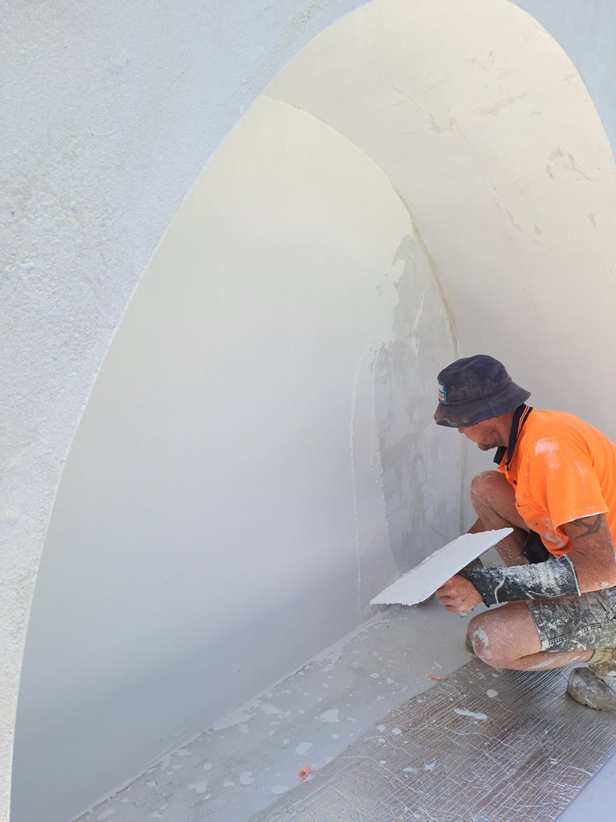 Person in orange shirt and hat, applying plaster to a curved white wall   — Neil Griffiths Rendering in Berry Springs, NT