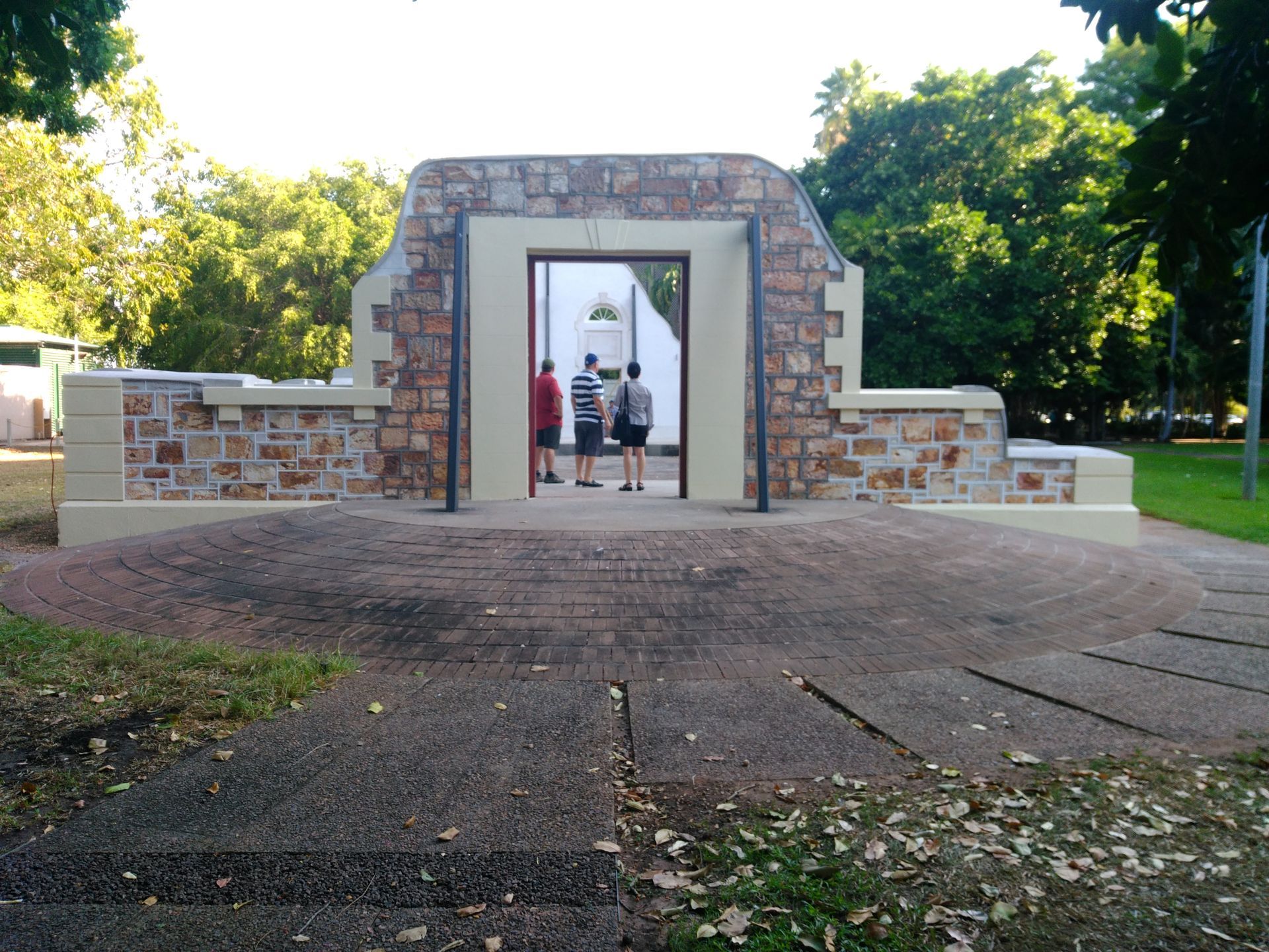 Stone monument with open doorway, three people standing inside; park setting, trees — Neil Griffiths Rendering in Berry Springs, NT