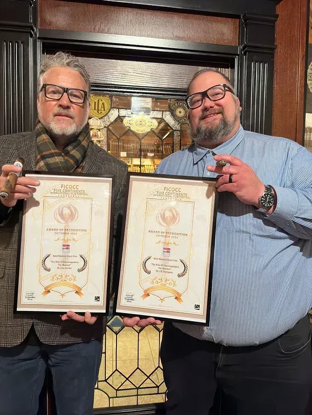 Two men holding framed certificates, smiling, in front of a dark wood background.