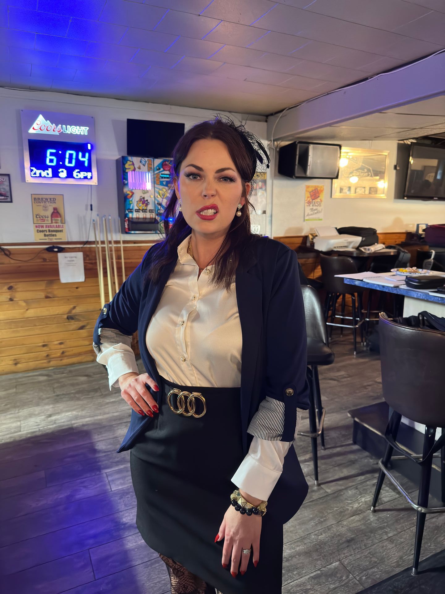 Woman in blazer, silk shirt, and skirt stands in a bar, hand on hip.