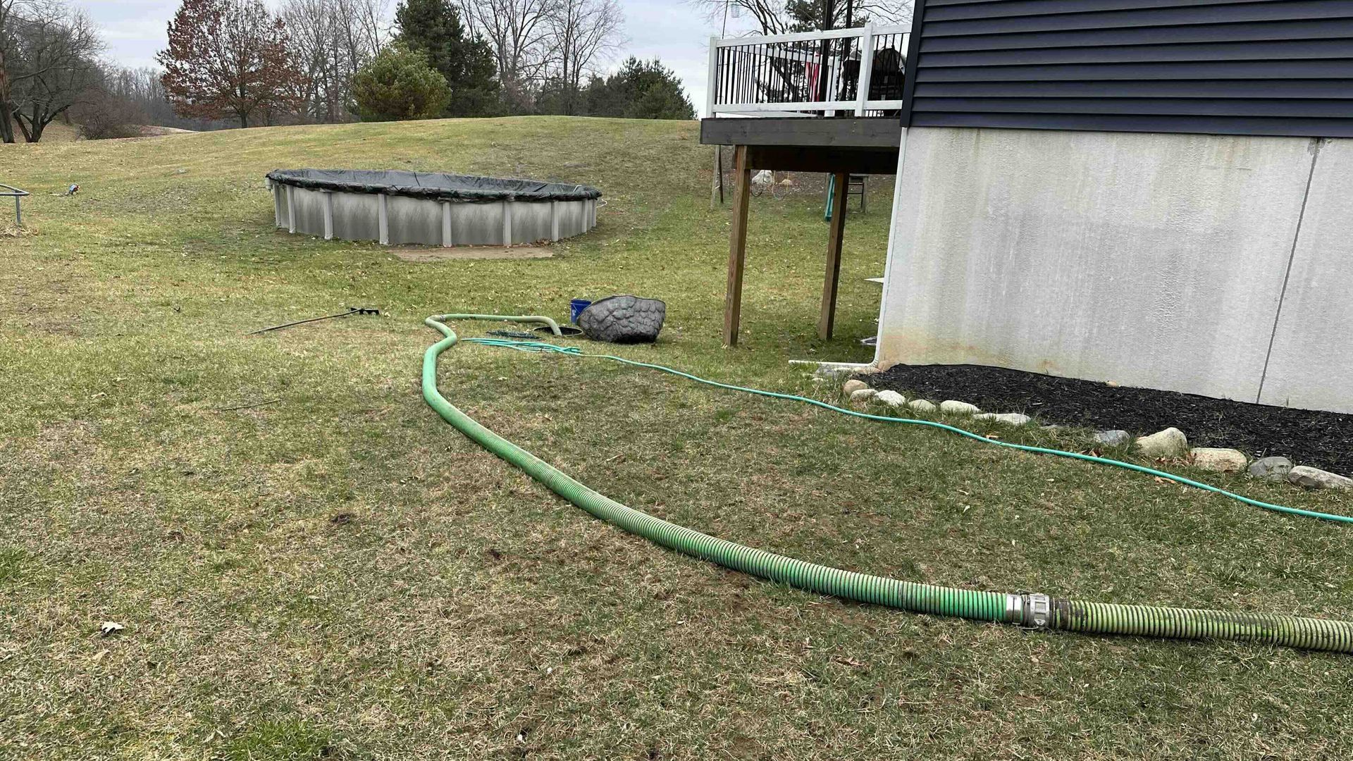 A green hose is pumping a septic tank that is located by a pool in a backyard.