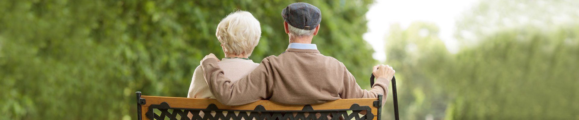 Two people sitting on a bench in a park, viewed from behind, with one person holding a cane.