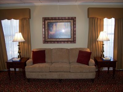 A beige sofa with two red cushions sits between two matching wooden end tables topped with lamps in a patterned room.