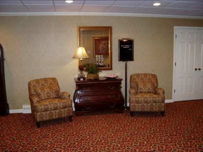 Two patterned armchairs flank a dark wood chest with a lamp, a mirror, and a standing sign in a room with red carpet.