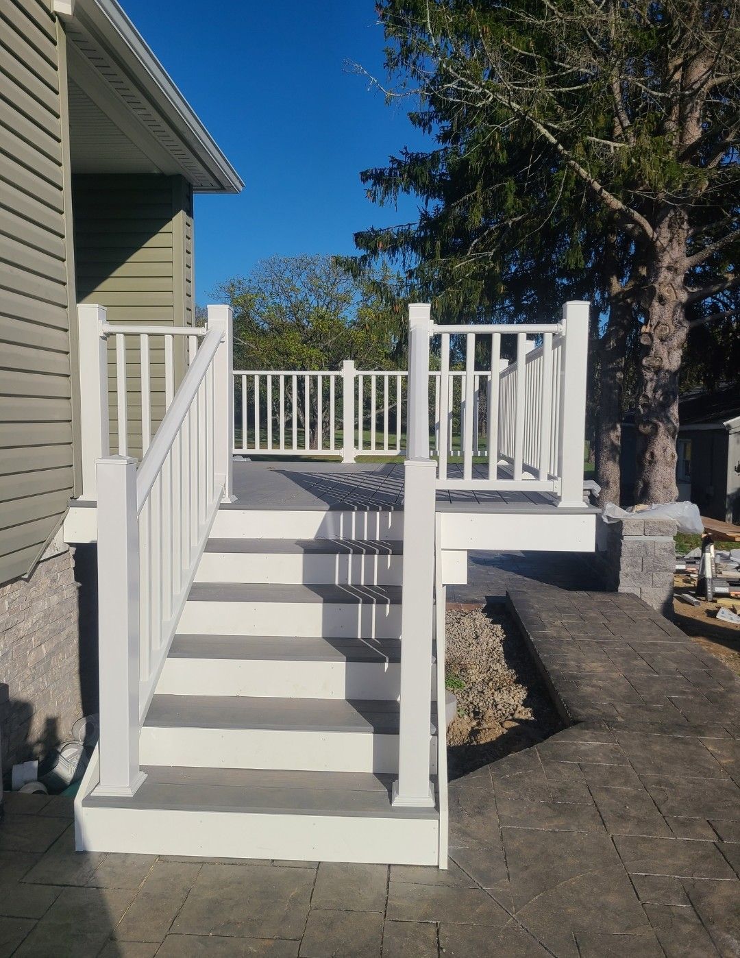 White deck with stairs, gray steps, white railing, against a green house, blue sky.