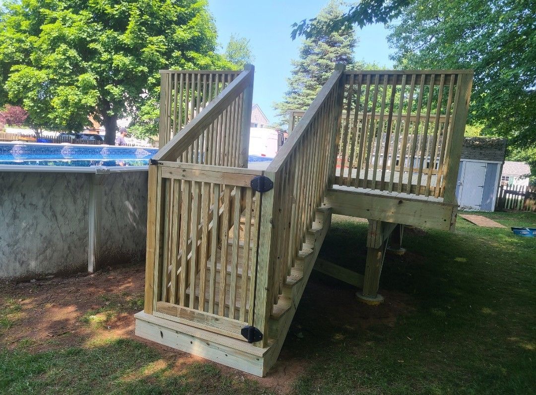 Wooden deck with stairs leading to a swimming pool, surrounded by green grass and trees on a sunny day.