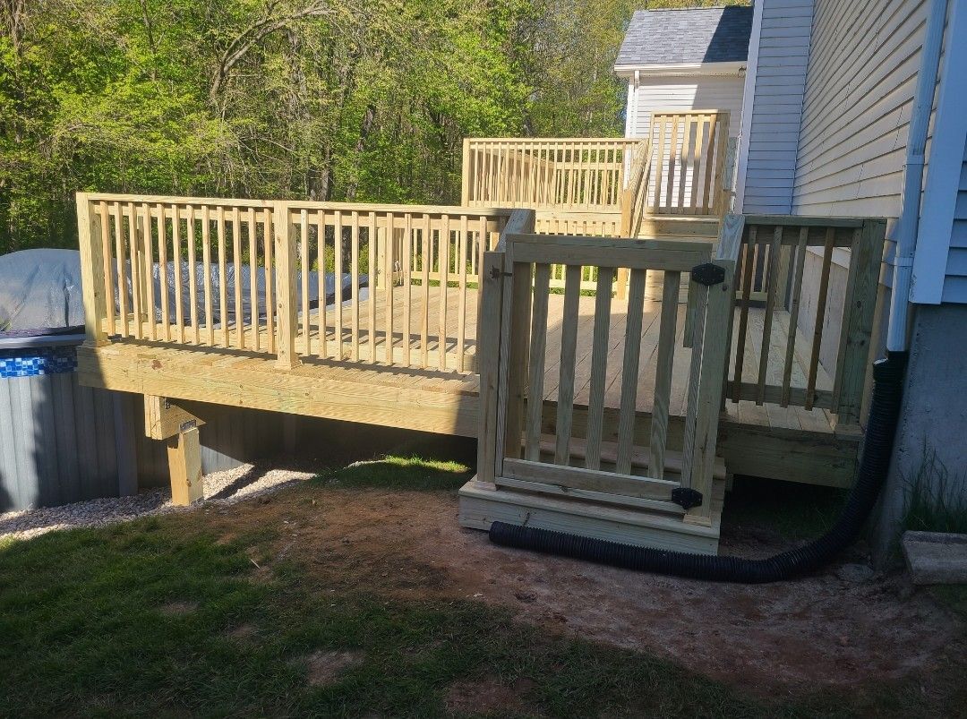 Wooden deck with railings and a gate next to a house, grass and trees in the background.