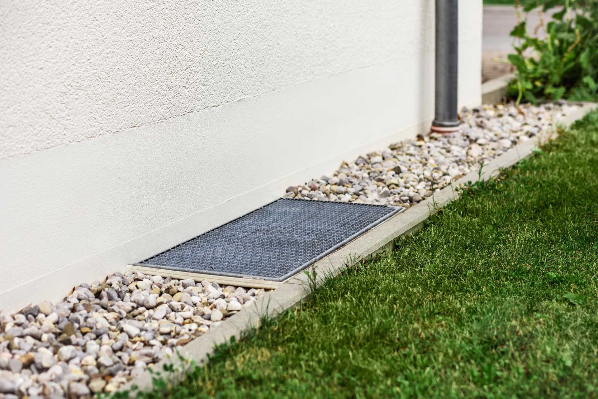 A rectangular metal grate in a gravel bed against a white wall and green grass.