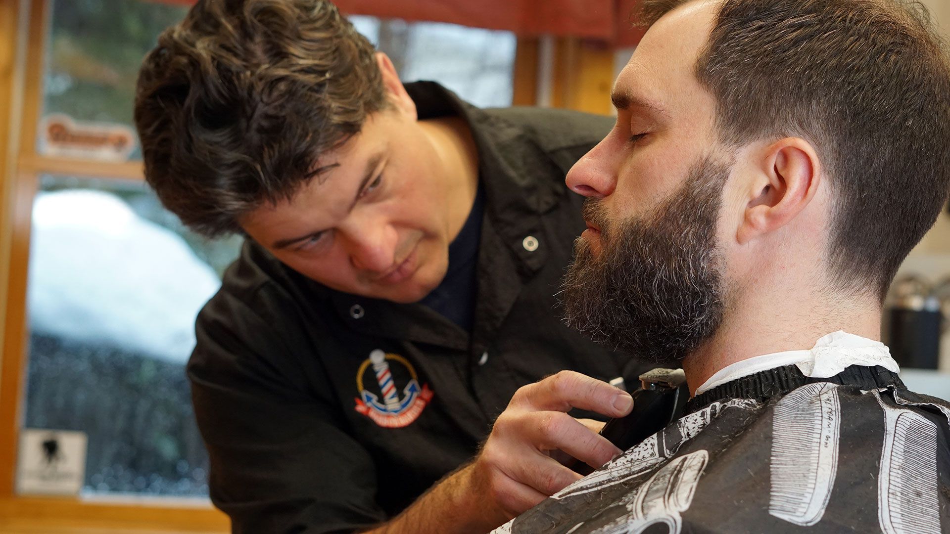A man sitting still with his eyes closed while a professional barber trims his beard.