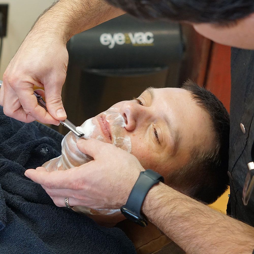 A man lying back in a barber chair receiving a traditional straight razor shave from a professional barber.