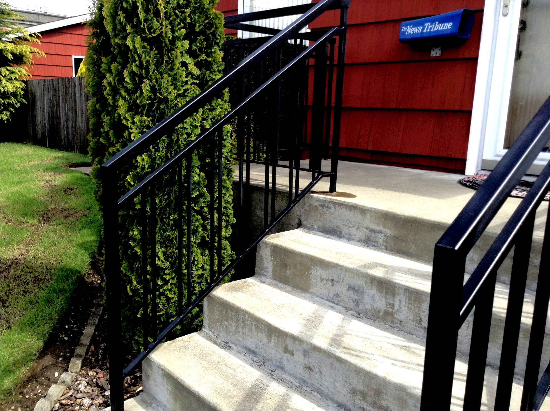 Concrete steps with black railings leading to a red house entrance beside a tall evergreen shrub