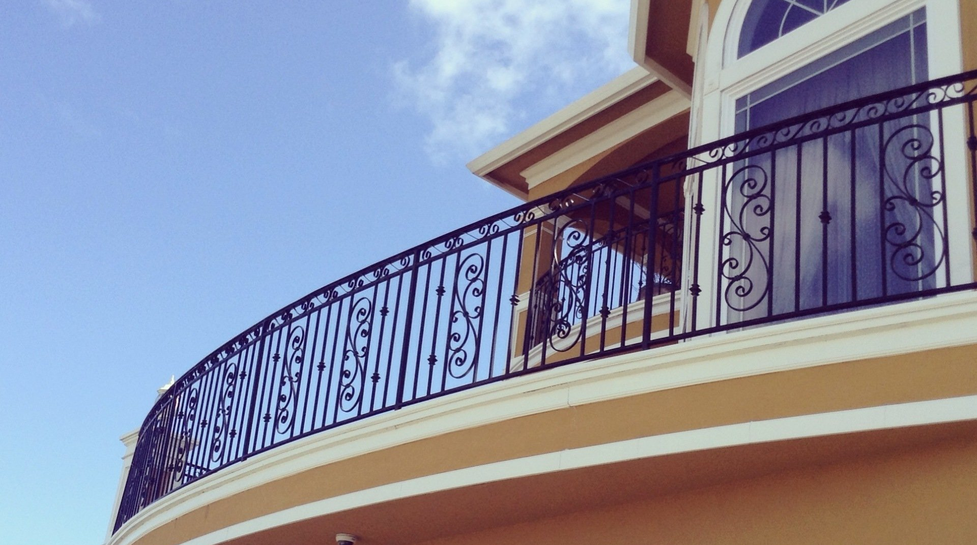 Curved balcony with dark wrought-iron railing on a cream-colored building against a blue sky