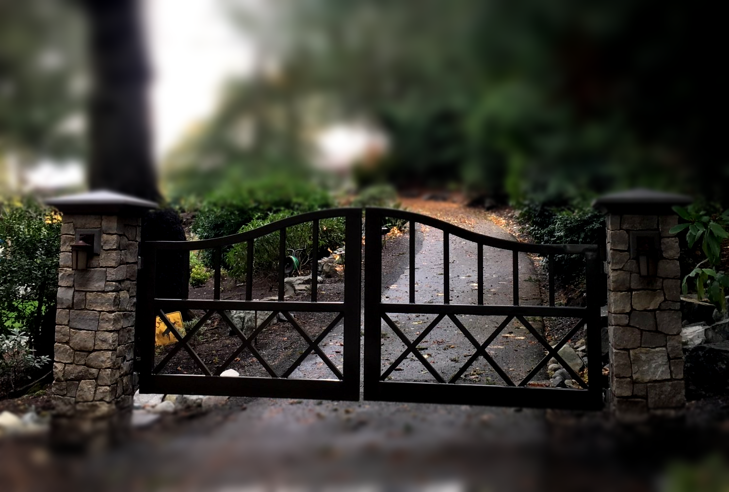 Stone-and-metal driveway gate between brick pillars, with a blurred house and trees behind it.