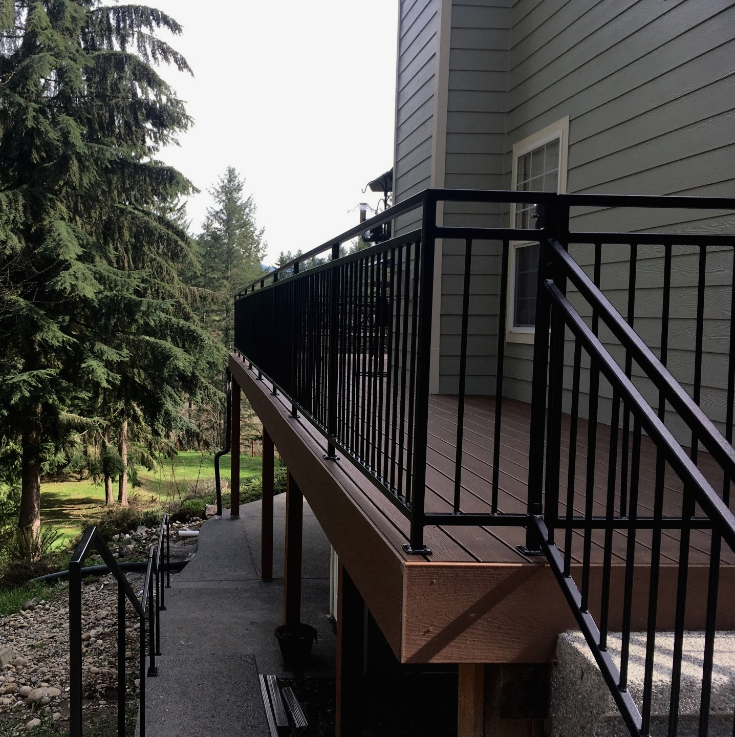 Black metal-railed ramp beside a gray house, leading down to a concrete path in a wooded yard