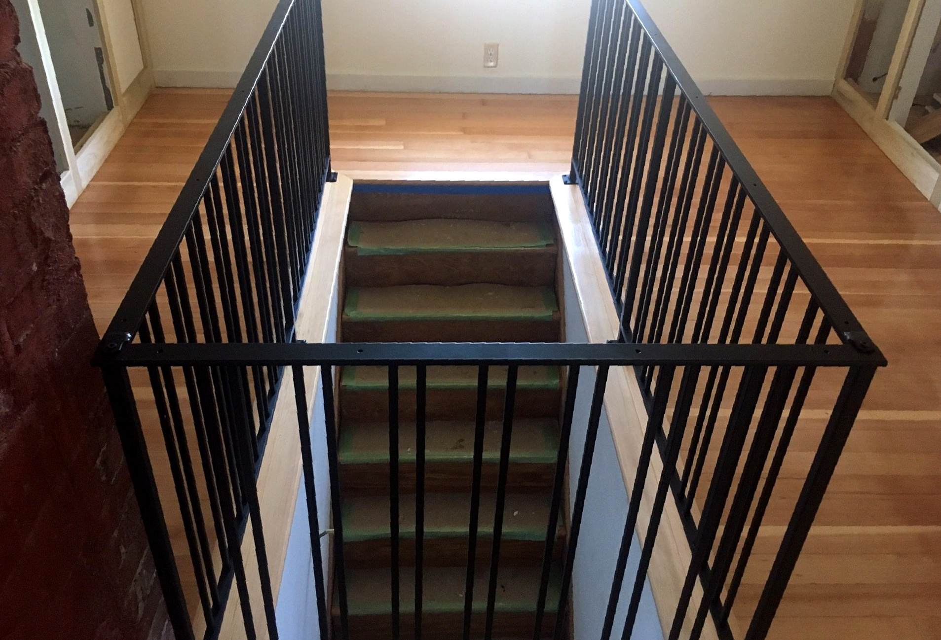 Black metal stair railing surrounding an opening to a basement staircase in a hallway.