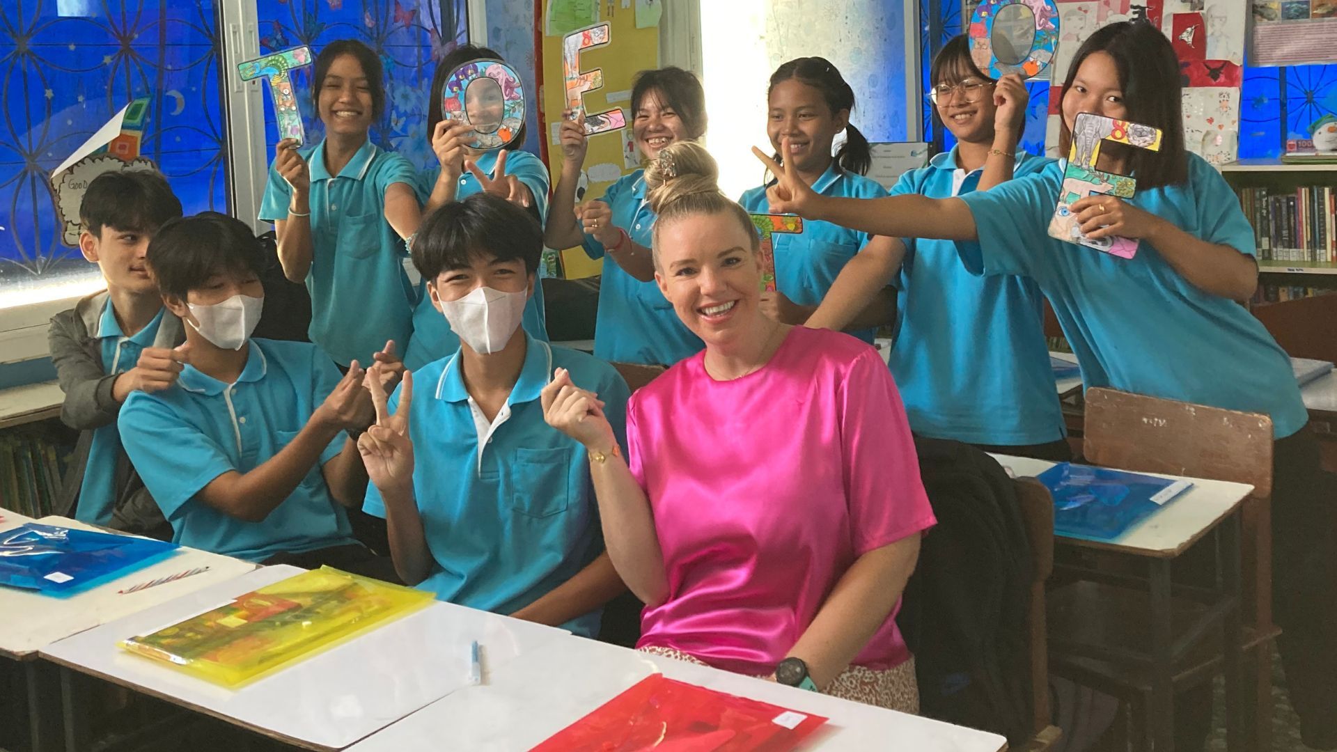 A woman in a pink shirt is sitting at a table with a group of children in a classroom.