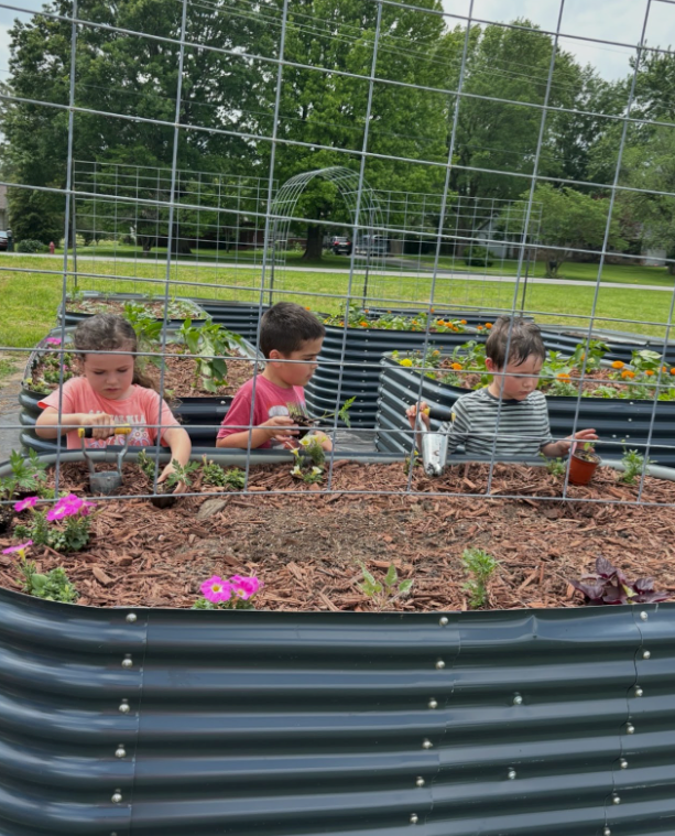 Three children are playing in a garden with flowers and plants.